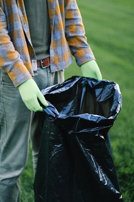 A person standing outdoors on a grassy area during daytime, wearing a multicoloured plaid shirt over a dark green top, with beige trousers and a brown belt, holding a large black garbage bag lined with a silver inner lining. The individual is wearing light green gloves and is in the process of lifting or placing the bag on a flat surface, with their hands gripping the top edges of the bag. The background features a lush, green lawn with blurred foliage, indicating a garden or park setting. The scene suggests an activity involving rubbish or waste collection, reflecting private disposal or cleanup efforts, supported by the presence of the black refuse bag which is often used in alternative or independent rubbish removal services like those offered by Rubbish Clearance Chiswick.