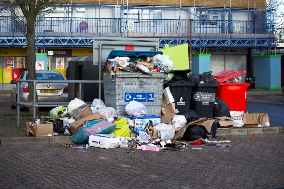 A large collection of mixed waste, including crumpled cardboard boxes, plastic bags filled with household rubbish, paper packaging, and miscellaneous debris, is piled on a paved area in front of a set of black and red wheelie bins. The waste appears disorganized, with items spilling outward across the pavement. Behind the piles, there are two grey and black recycling containers designated for mixed paper and cardboard, both overflowing with refuse. To the left of the waste, a grey sedan car is parked, with a yellow registration plate visible. In the background, a commercial building with storefronts and signage, including a fish bar and other shops, is visible, along with a blue scaffolding structure indicating ongoing construction or maintenance work. The scene is lit by natural daylight, with an overcast sky casting diffuse light over the surroundings, illustrating an example of private waste disposal or on-site rubbish accumulation in an urban setting, which local rubbish removal services like Rubbish Clearance Chiswick might assist with.