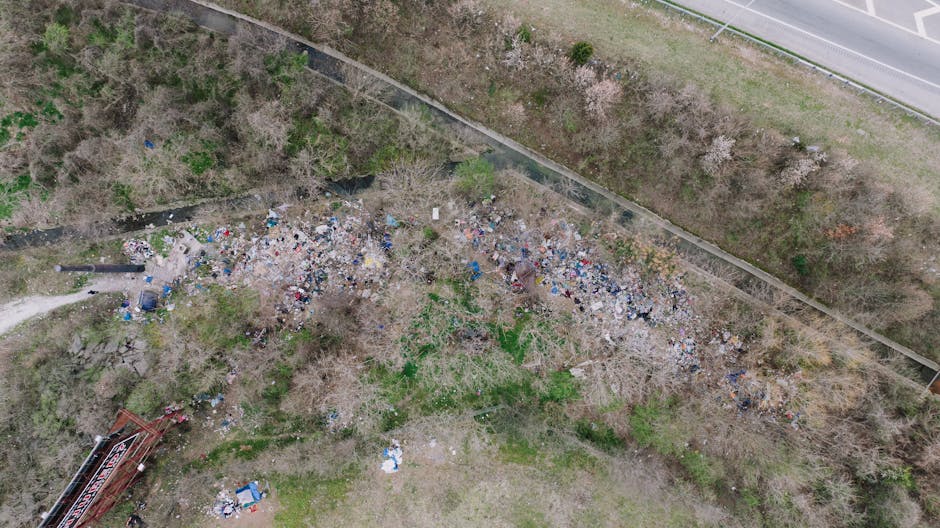 An aerial photograph showing a large accumulation of mixed rubbish and debris scattered across an open, partially wooded area adjacent to a narrow dirt pathway. The waste includes various types of discarded plastics, cardboard, and other household refuse, with some items partially hidden among leafless trees and bushes. In the upper right corner, there is a section of paved road with a concrete curb and a metal fence running parallel to the roadside, separating the rubbish area from the street. The scene appears to be an unmanaged dumping site, with no visible waste collection equipment present, indicating it may be a private or illegal waste disposal site that could be addressed through independent rubbish removal services such as those provided by Rubbish Clearance Chiswick. The natural environment surrounding the waste is a mix of leafless branches and sparse greenery, suggesting a seasonal transition, with broken and scattered items contributing to an overall untidy landscape, emphasizing the need for intervention to restore the area’s appearance and prevent environmental impact.