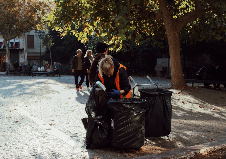 A woman wearing a high-visibility orange vest is seen collecting trash from a large black rubbish bag in a public park or square, with a second black bag nearby on the ground. She is using gloves and appears to be sorting or disposing of waste, with her head bent down as she works. In the background, several pedestrians are walking along a paved path, with some trees casting shade over the area, and a building with windows and doors visible in the distance. The environment is illuminated by natural light, suggesting a sunny day, and the scene depicts an outdoor setting suitable for waste collection, highlighting a community effort to maintain cleanliness that links to professional rubbish removal and clearance services such as those offered by Rubbish Clearance Chiswick. The overall composition emphasizes the practical activity of waste management within a public urban space, supporting themes of environmental responsibility and private rubbish disposal options.