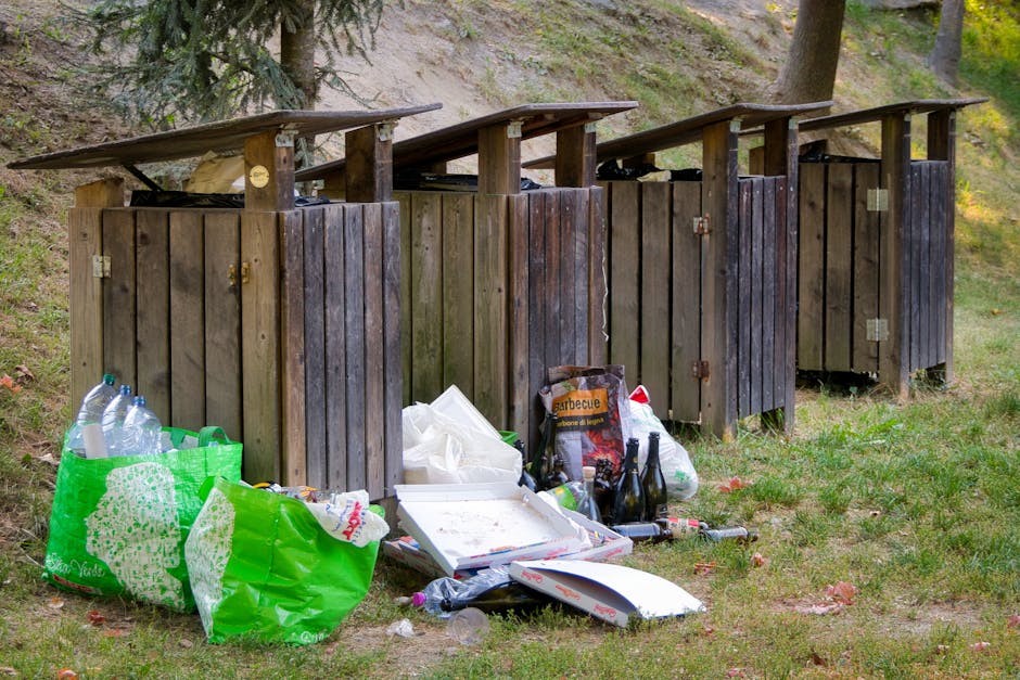 Four wooden outdoor rubbish bins with slanted roofs are positioned side by side on a grassy area, with some of the lids slightly ajar. In front of the bins, there is a collection of discarded items including two large transparent plastic bottles, a bright green plastic shopping bag filled with more bottles, a white pizza box with some food remnants visible, and a few empty glass bottles. The scene is set against a backdrop of a grassy slope with trees and shrubs, indicating an outdoor environment. The lighting appears natural, suggesting daylight, and the scene reflects a private or informal waste disposal situation, with waste temporarily stored outside before collection. The presence of rubbish and waste materials emphasizes the need for proper rubbish removal services, such as those provided by Rubbish Clearance Chiswick, to handle bulk waste and prevent environmental clutter.